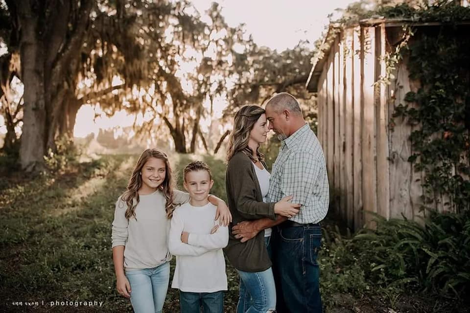 family portrait of mom, dad, son, and daughter next to barn under oak trees at sunset